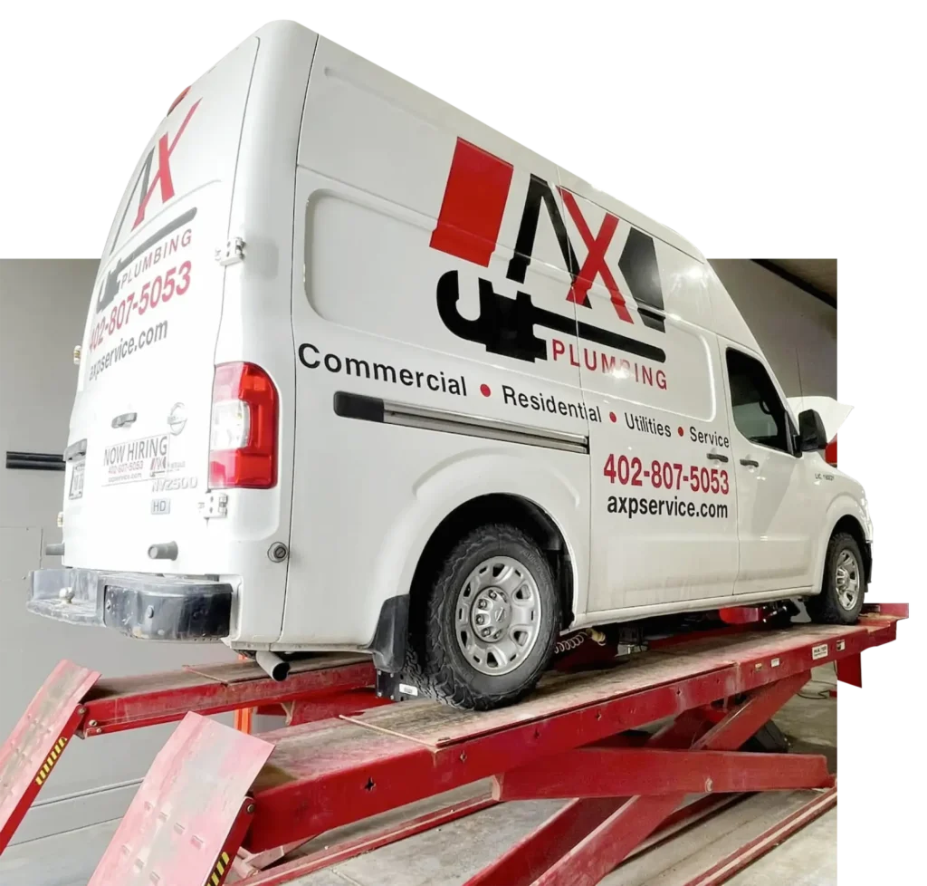 A white AX Plumbing service van is elevated on a red hydraulic lift. The van displays company branding, phone number, and lists services: commercial, residential, utilities, and service.