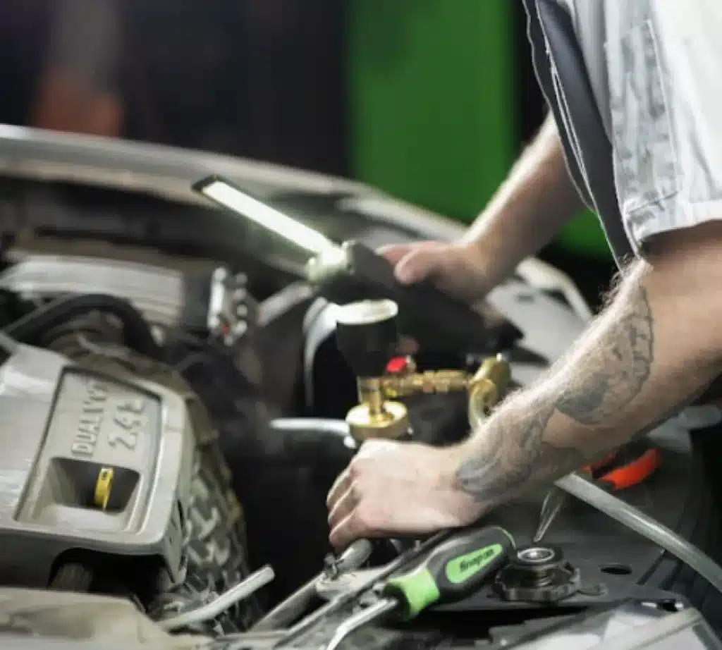 A mechanic with tattoos on his arms works under the hood of a car, using tools and a bright work light to inspect the engine while performing auto air conditioning repairs.