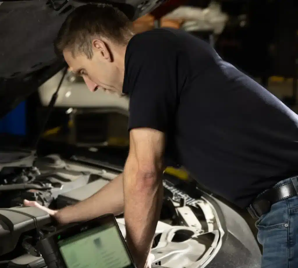 A mechanic in a dark polo shirt leans over a car engine, inspecting it with a diagnostic tool on a nearby laptop. The hood is open in the garage, highlighting the hands-on expertise needed in auto repair.