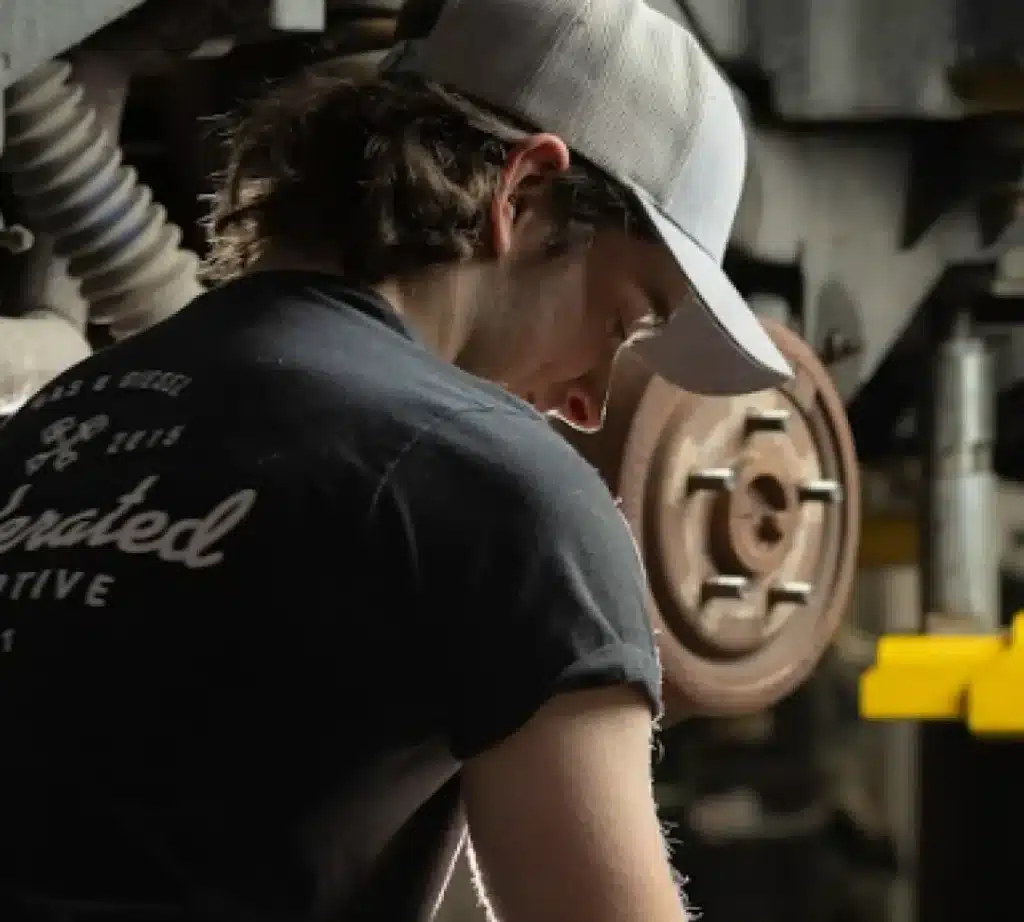 A person wearing a gray cap and black T-shirt works on a car, focusing on brake repair with the brake disc and mechanical parts visible in the background. The person's face is partially turned away from the camera.