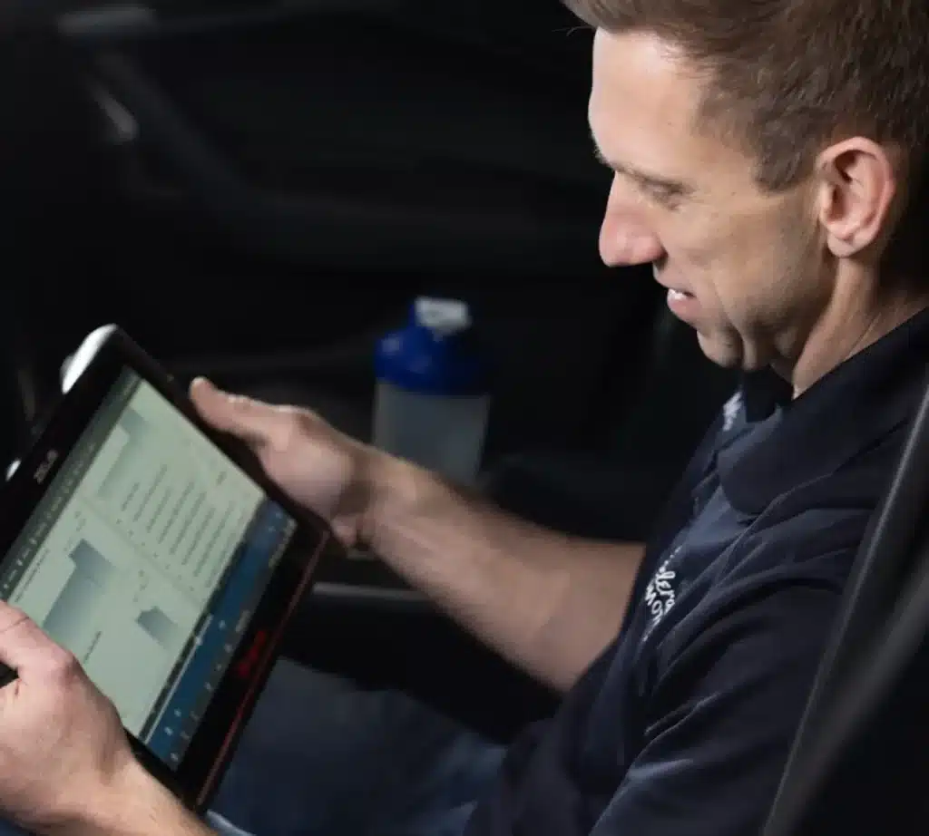 A man sitting in a car looks at a tablet displaying charts, data, and engine diagnostic information. A blue water bottle is visible in the background.