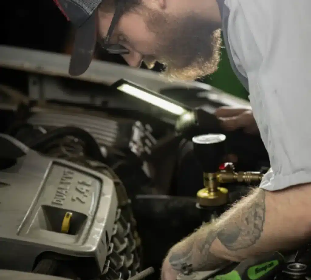 A bearded mechanic wearing a cap and glasses performs a vehicle inspection on a car engine, using a diagnostic tool and flashlight. His arm displays a visible tattoo.