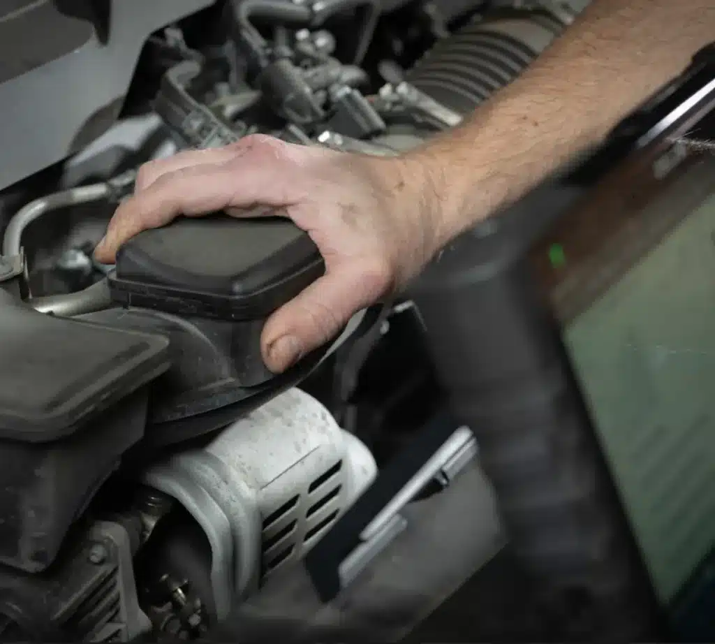 A close-up of a mechanic’s hand inspecting or repairing a car engine for preventative maintenance, with parts visible. The person is using a laptop nearby, possibly for diagnostics.