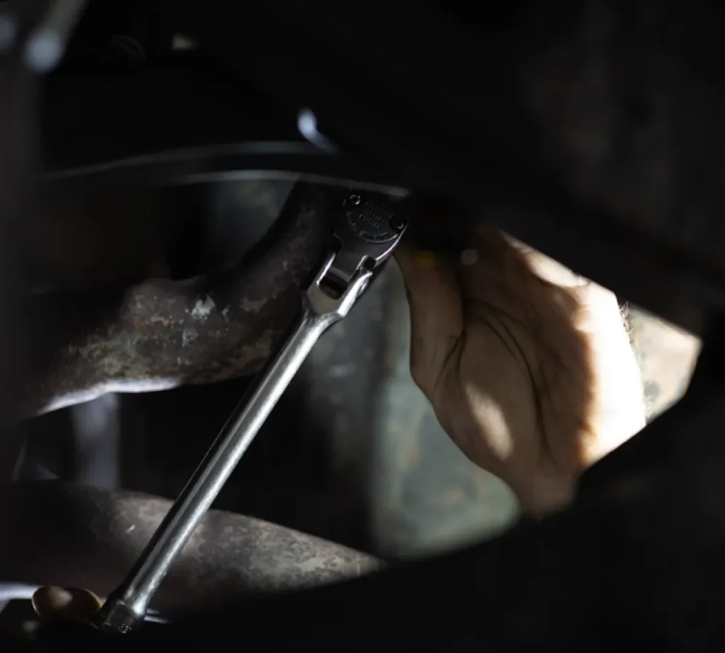 A close-up of a person's hand using a ratchet wrench to tighten a bolt on a metal pipe, likely performing steering or suspension repair on a car engine or machinery in low light.