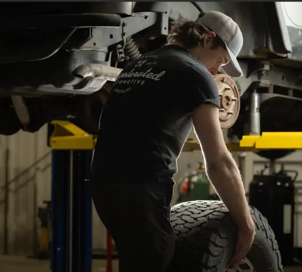 A mechanic wearing a cap and black t-shirt lifts a large tire near a vehicle raised for alignment on a hydraulic lift in an auto repair shop.