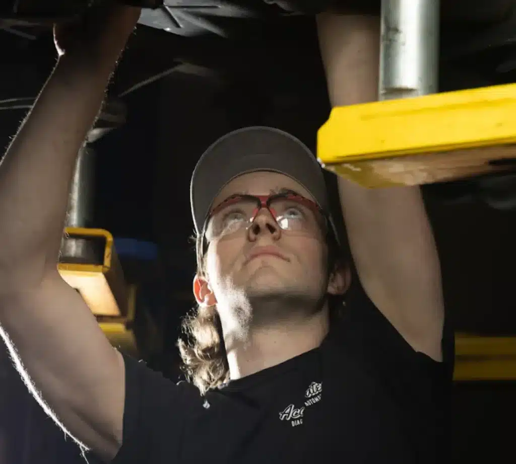 A person wearing safety glasses and a cap is working underneath a vehicle, reaching up with both arms, likely performing transmission maintenance or repairs in a well-lit workshop.