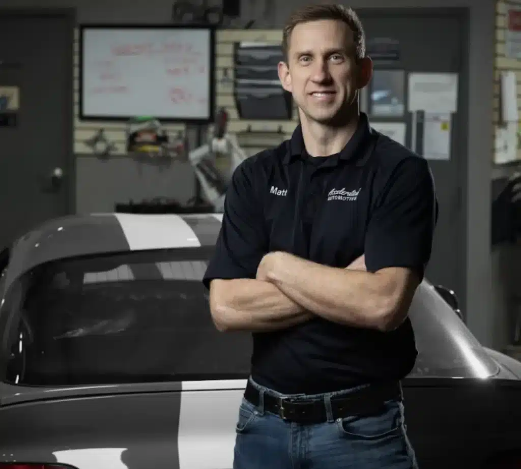 A man in a dark polo shirt stands with arms crossed in front of a silver car in an auto repair workshop, with vehicles, shelves, tools, and a whiteboard visible in the background.
