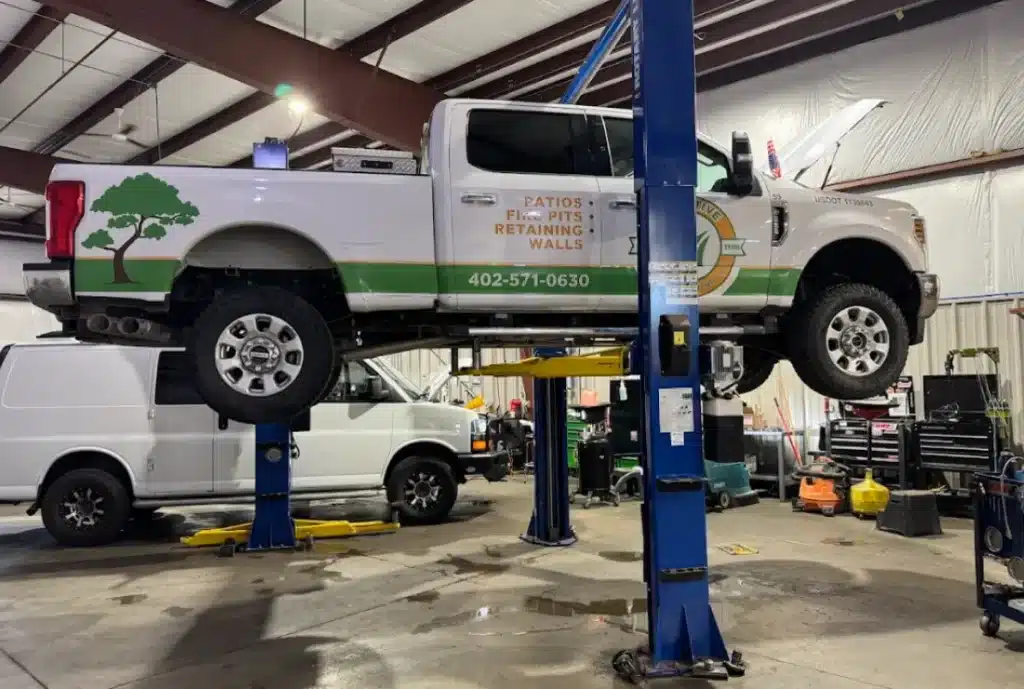 A white fleet pickup truck with landscaping company branding is elevated on a hydraulic lift in an auto repair shop. Tools, equipment, and another white van are visible in the background.